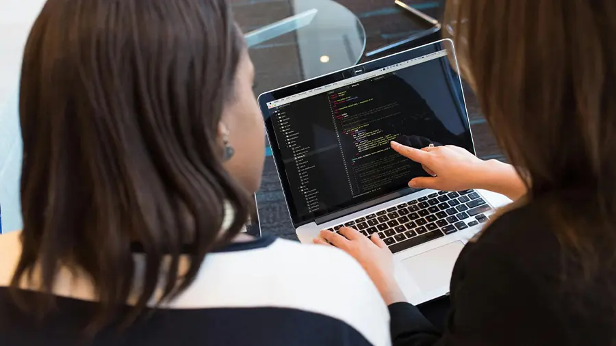 An image of one woman explaining, editing and pointing at code on a laptop. She is explaining it to the other woman sitting next to her.